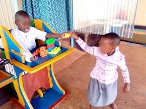 A child seated in a special chair with a tray, playing with another child